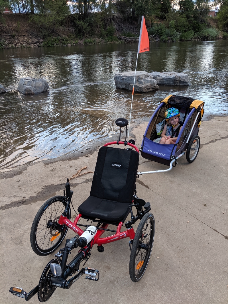 Catrike Trail and hitched trailer (occupied by a smiling kid) parked on the South Platte River bank.